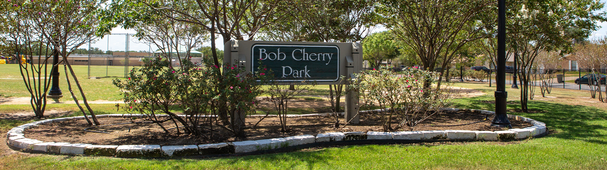 Bob Cherry Park sign in a garden bed with bushes and trees.